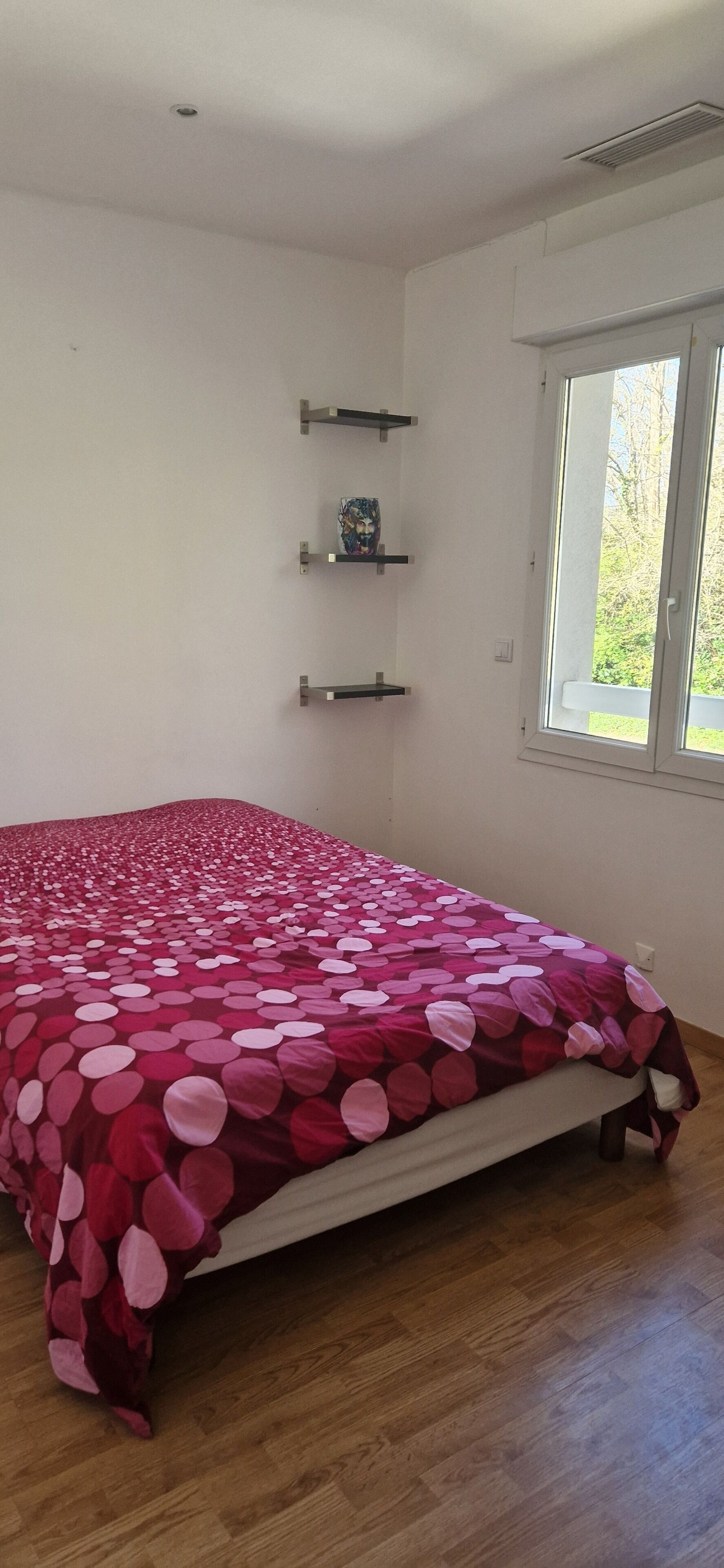 Een slaapkamer met een groot bed, houten vloeren, een raam met uitzicht op groen en drie wandplanken.