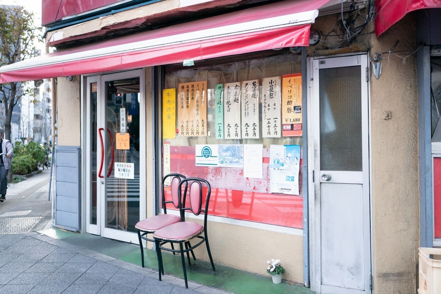 A restaurant with a red awning and a glass door. There are several menus displayed outside, and two chairs are placed in front of the entrance.