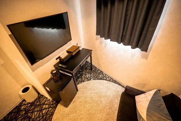 Une chambre d’hôtel dotée d’un téléviseur à écran plat fixé au mur, d’un petit bureau noir avec une machine à café, d’une poubelle et d’un lit avec une tête de lit sombre.