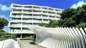 A modern multi-story hotel with balconies, a curved white structure in the foreground, and lush greenery.