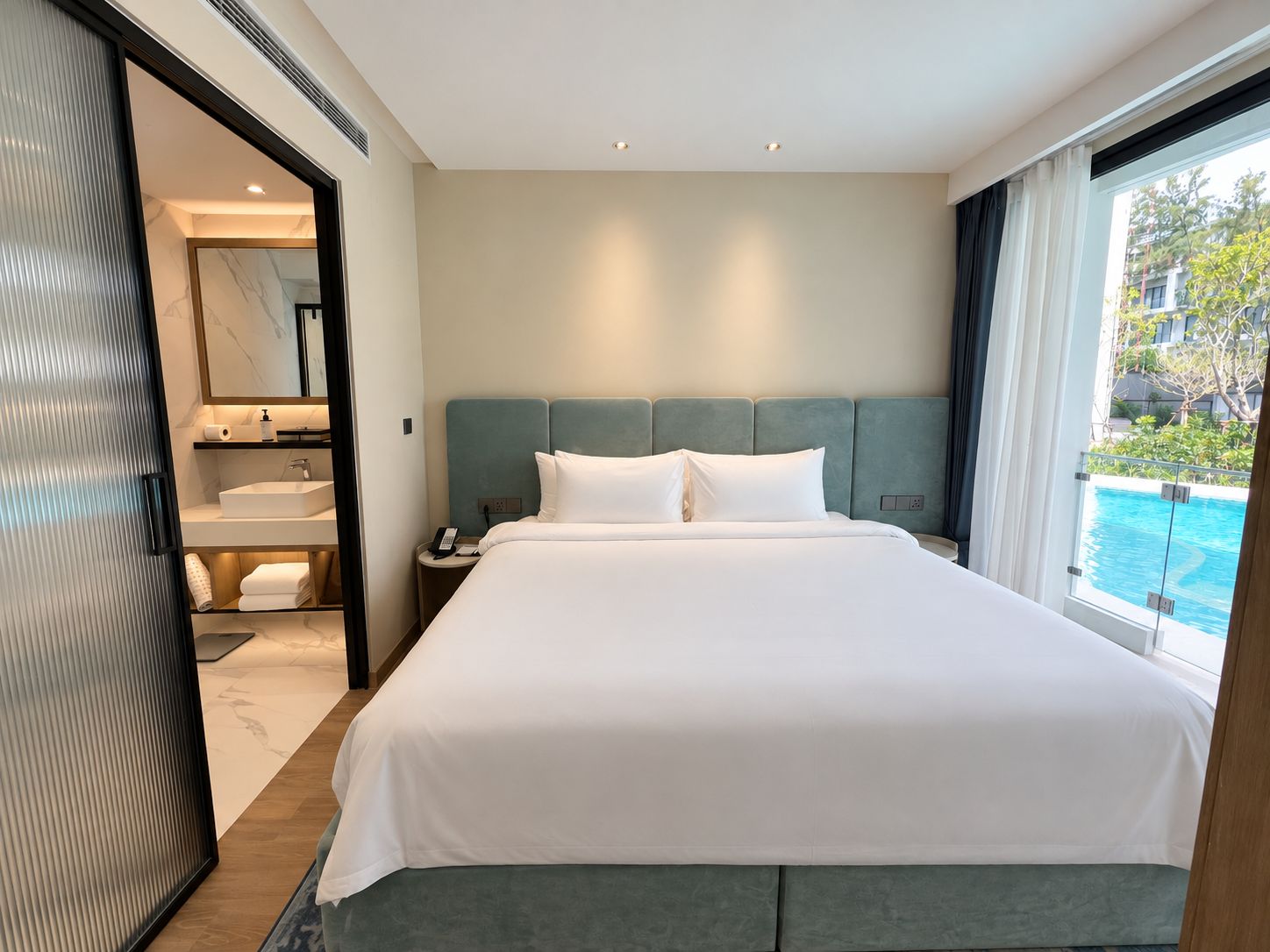 Una habitación de hotel con una cama grande, un baño con lavamanos y espejo, y vista a una piscina.