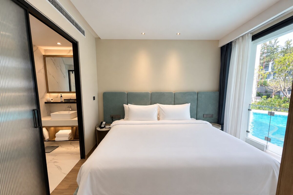 A hotel room with a large bed, a bathroom with a sink and mirror, and a view of a swimming pool.