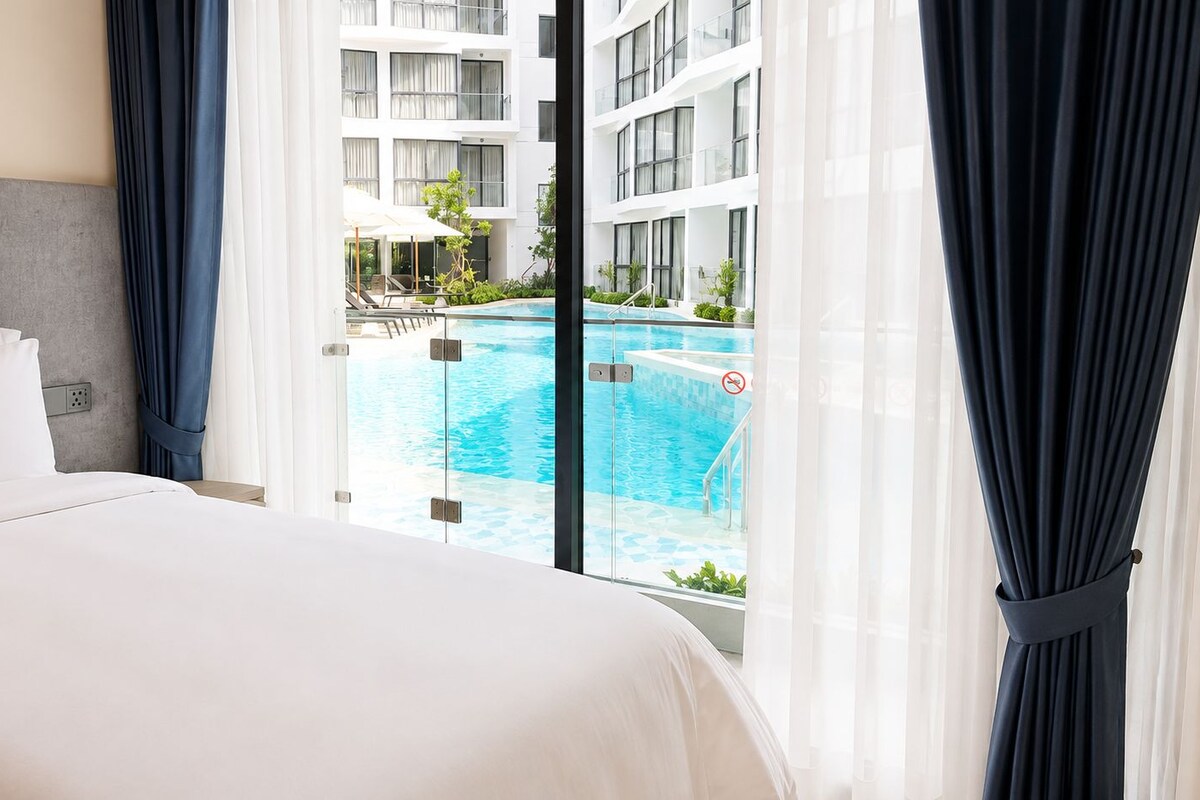 A hotel room with a bed, a view of a swimming pool, and a building in the background.