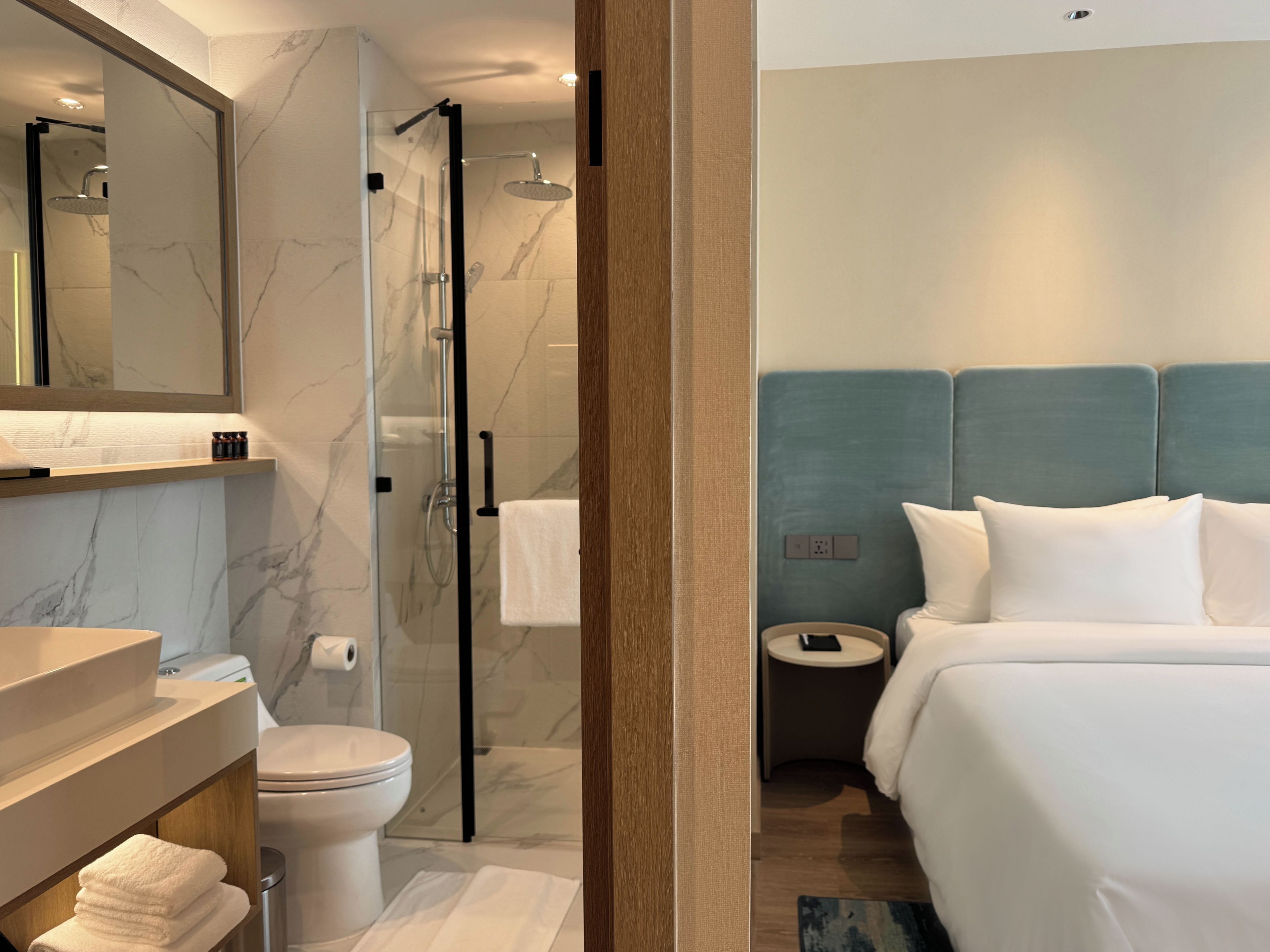 A hotel room with a bathroom including a toilet, sink, and shower. There is a bed with pillows and a bedside table.