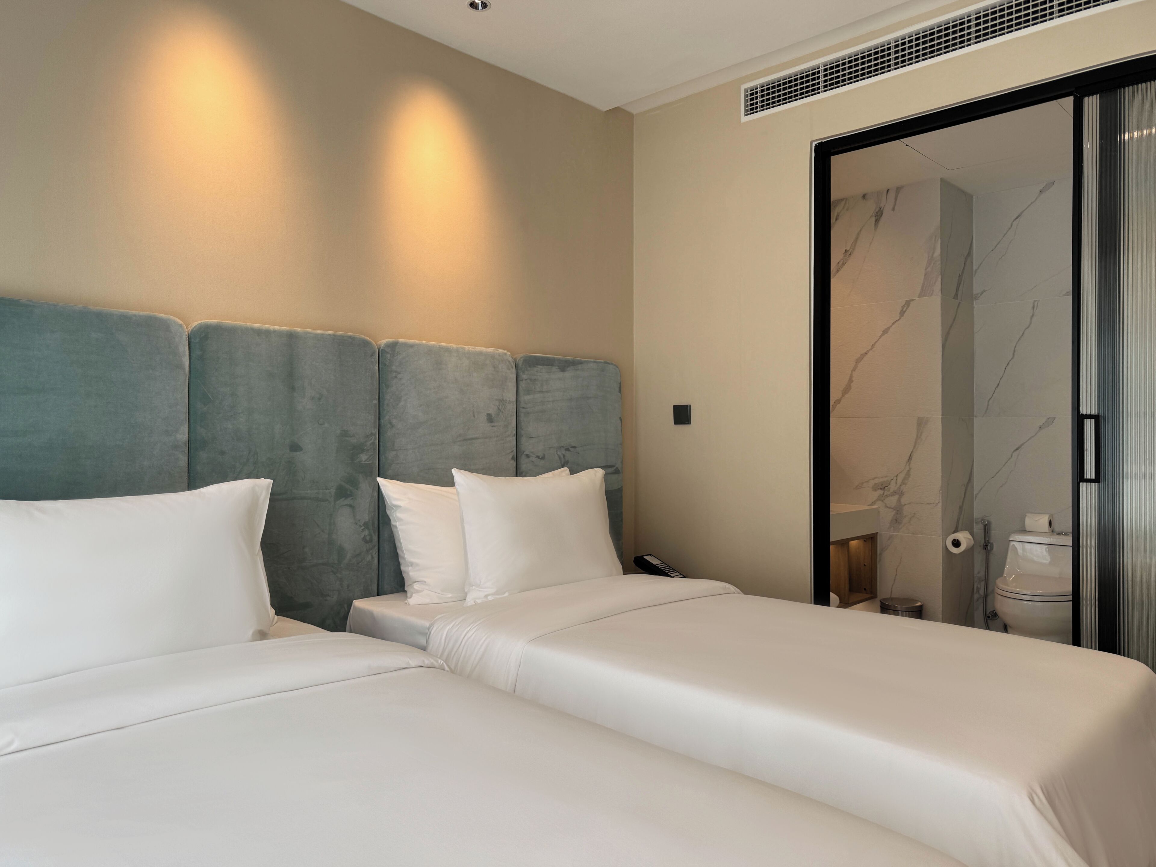 Habitación de hotel con dos camas, una pared de mármol y un baño visible a través de una puerta de vidrio.
