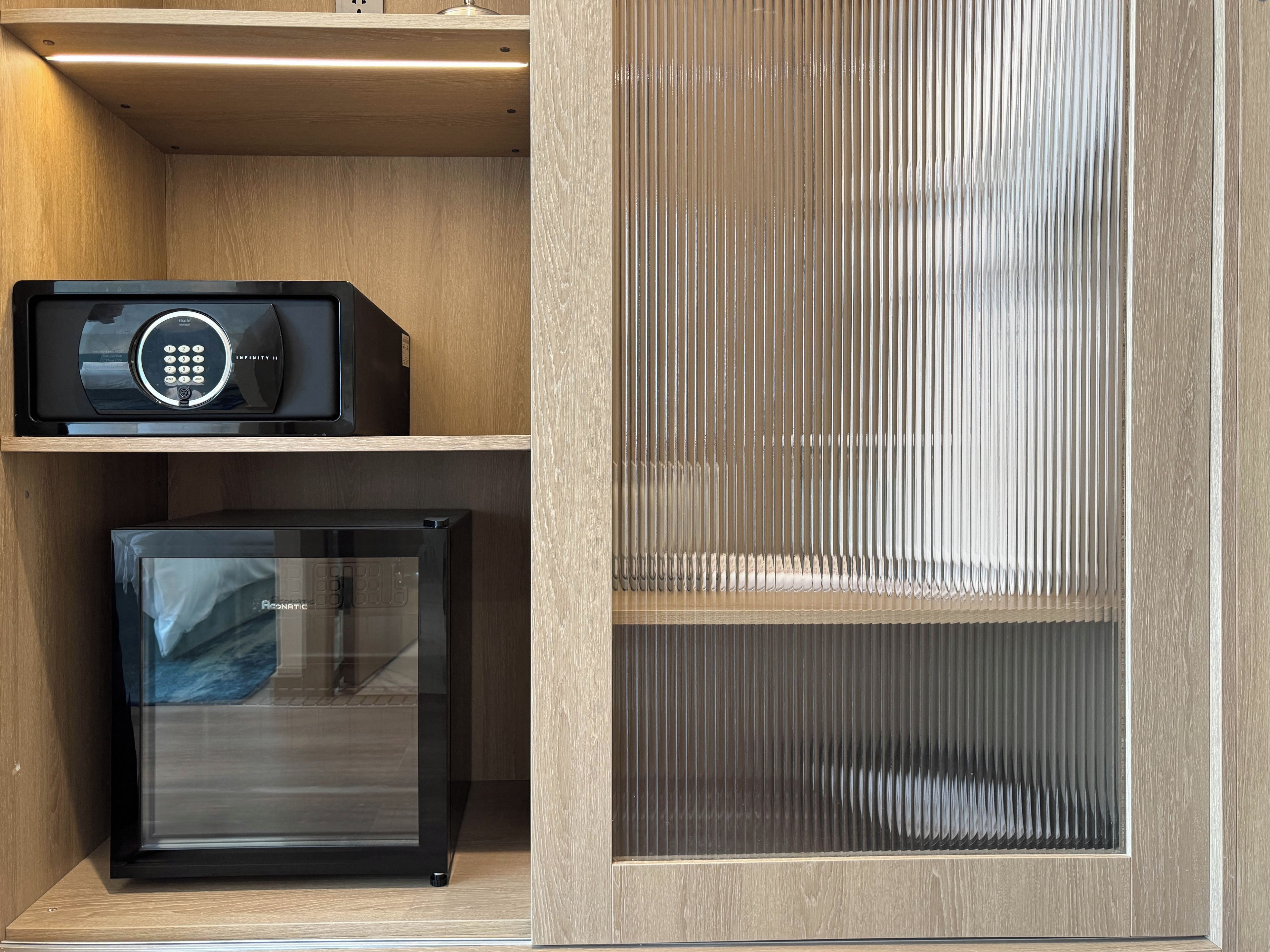 A compact room with a built-in refrigerator, a black electronic device on a shelf, and a wooden panel with vertical slats.