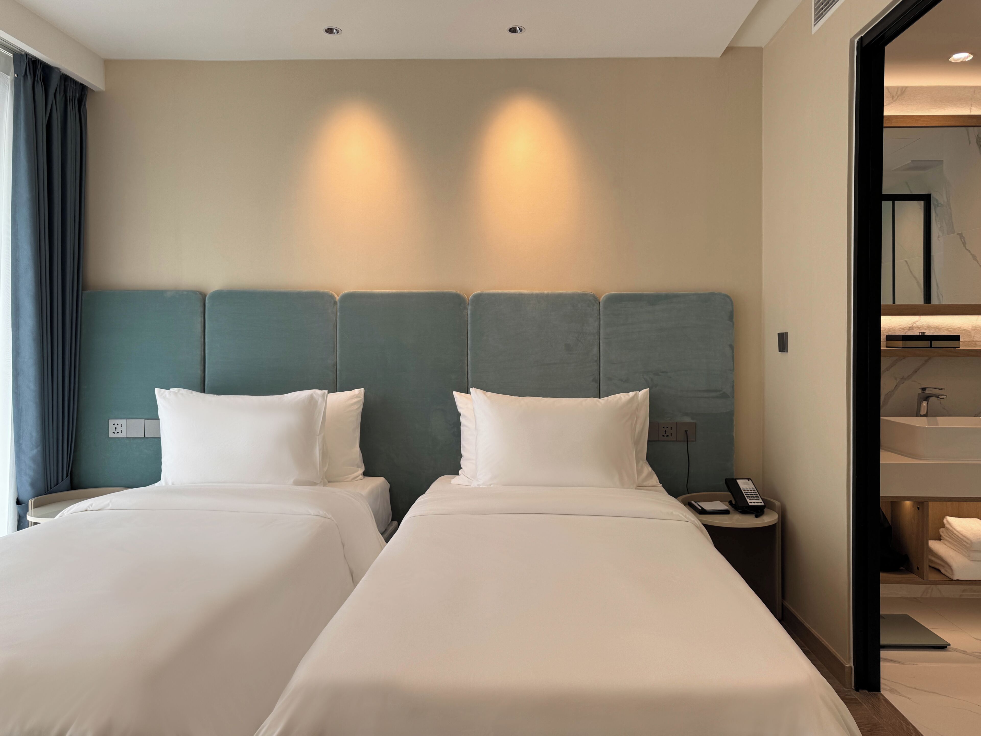 A hotel room with two beds, a nightstand with a phone, a bathroom with a sink and toilet, and a mirror.