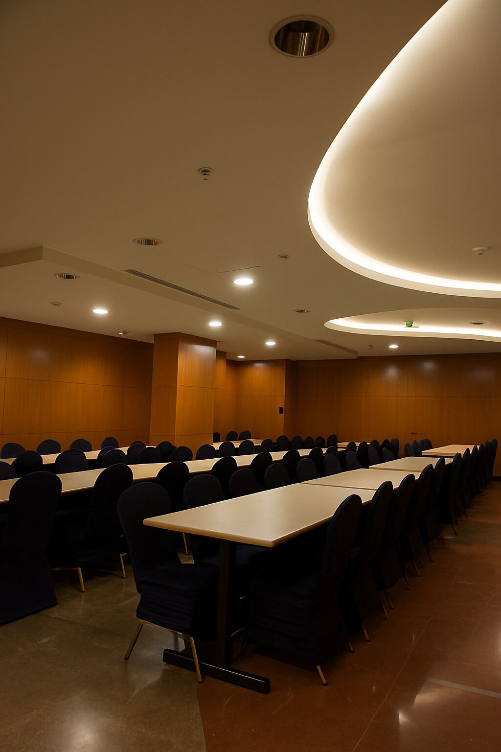 A conference room with rows of tables and chairs, recessed ceiling lighting, and wooden panel walls.