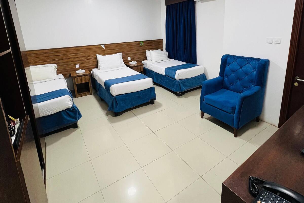 A hotel room with two beds, a blue armchair, a desk with a telephone, and an air conditioning unit on the wall.
