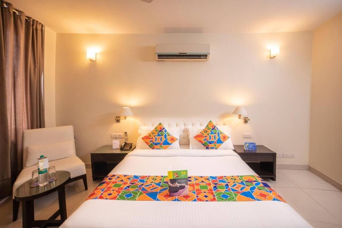 A hotel room with a bed, two nightstands, a chair, an air conditioner, and a window with curtains.