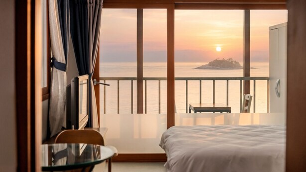 A room with a bed, a glass table, and a chair, with a balcony overlooking a sunset and a small island. - Sokcho SBS Pension (Sokcho)