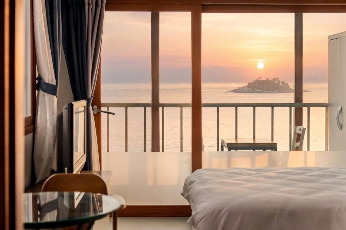 Una habitación con cama, una mesa de vidrio y una silla, con balcón que da a un atardecer y una pequeña isla.