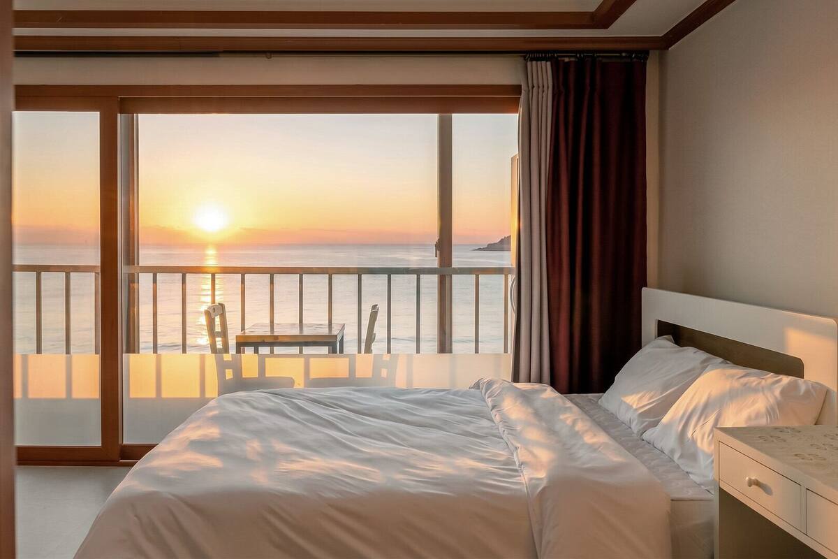 Una habitación de hotel con una cama, una mesita de noche, una silla y vista al atardecer sobre el océano.