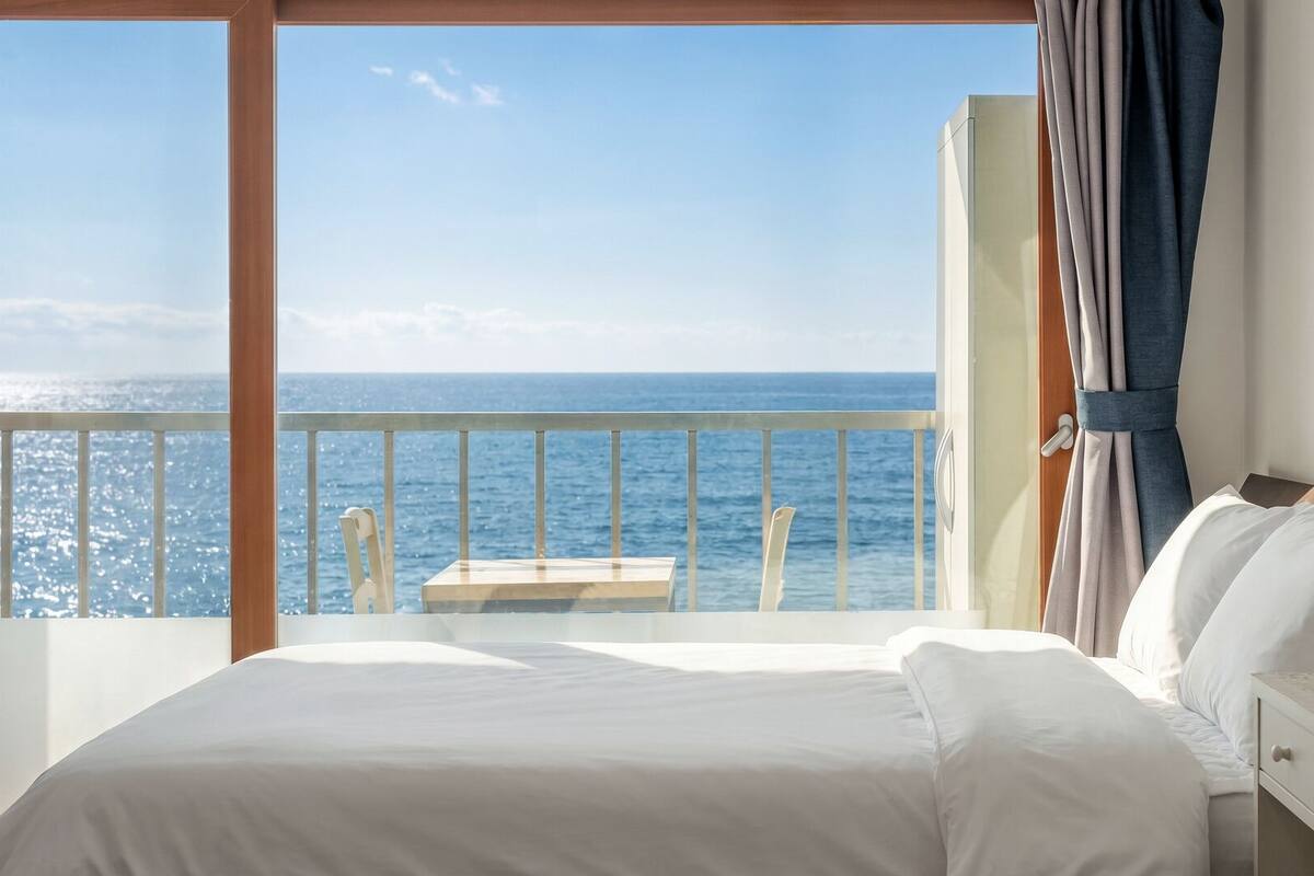 Habitación de hotel con un ventanal grande, una cama, una mesita de noche y vistas al mar.