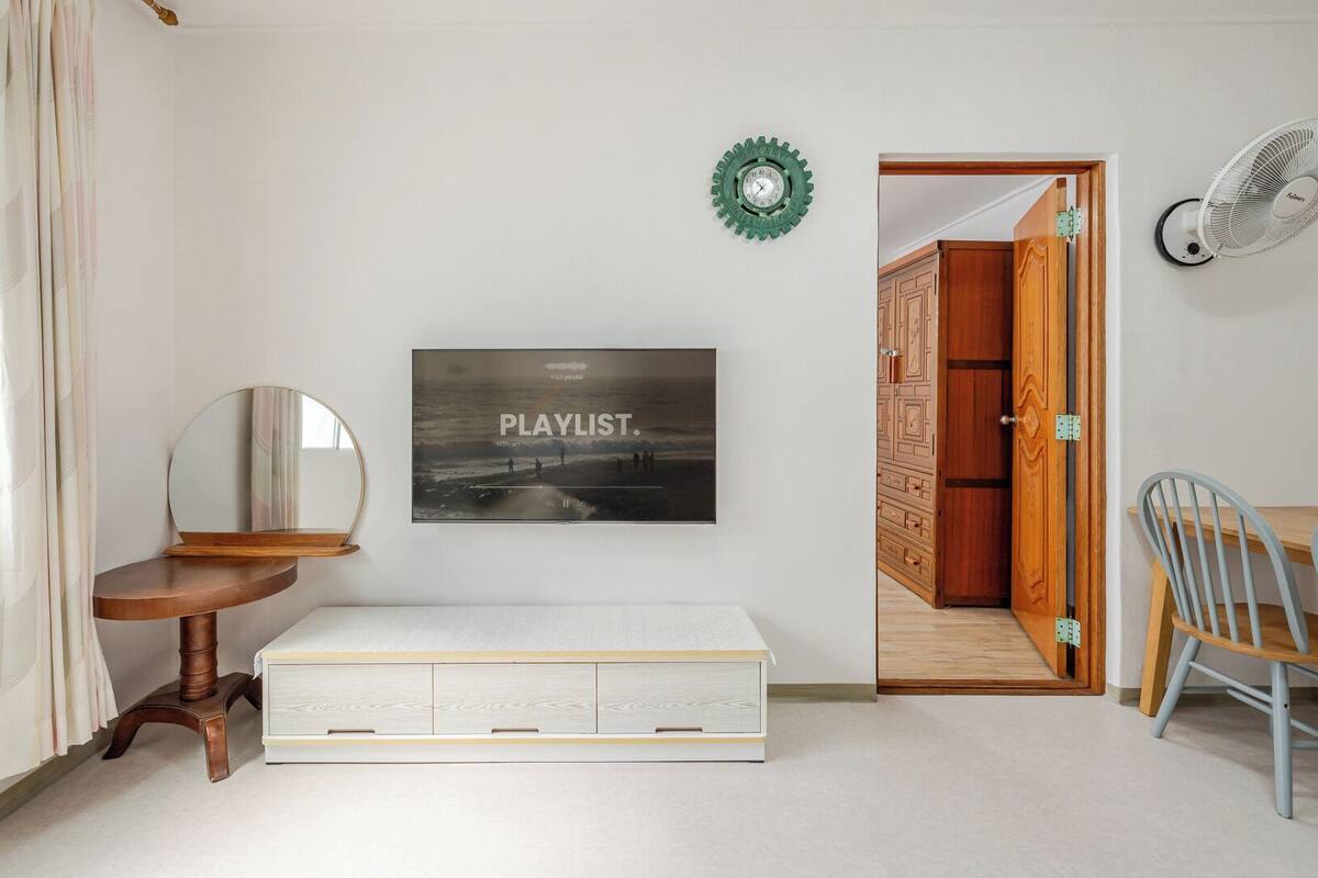 Una habitación con cama, espejo, reloj en la pared, puerta que da a otra habitación y ventilador fijado a la pared.