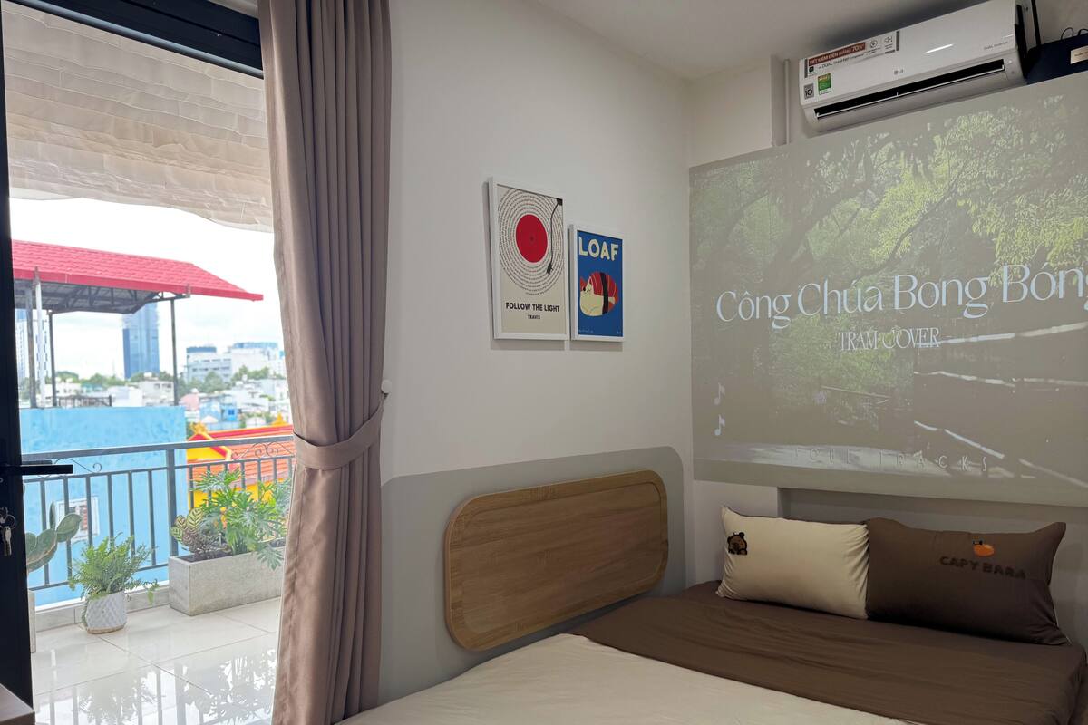 A hotel room with a bed, a projector screen displaying a travel show, a window with curtains, and a balcony with plants.