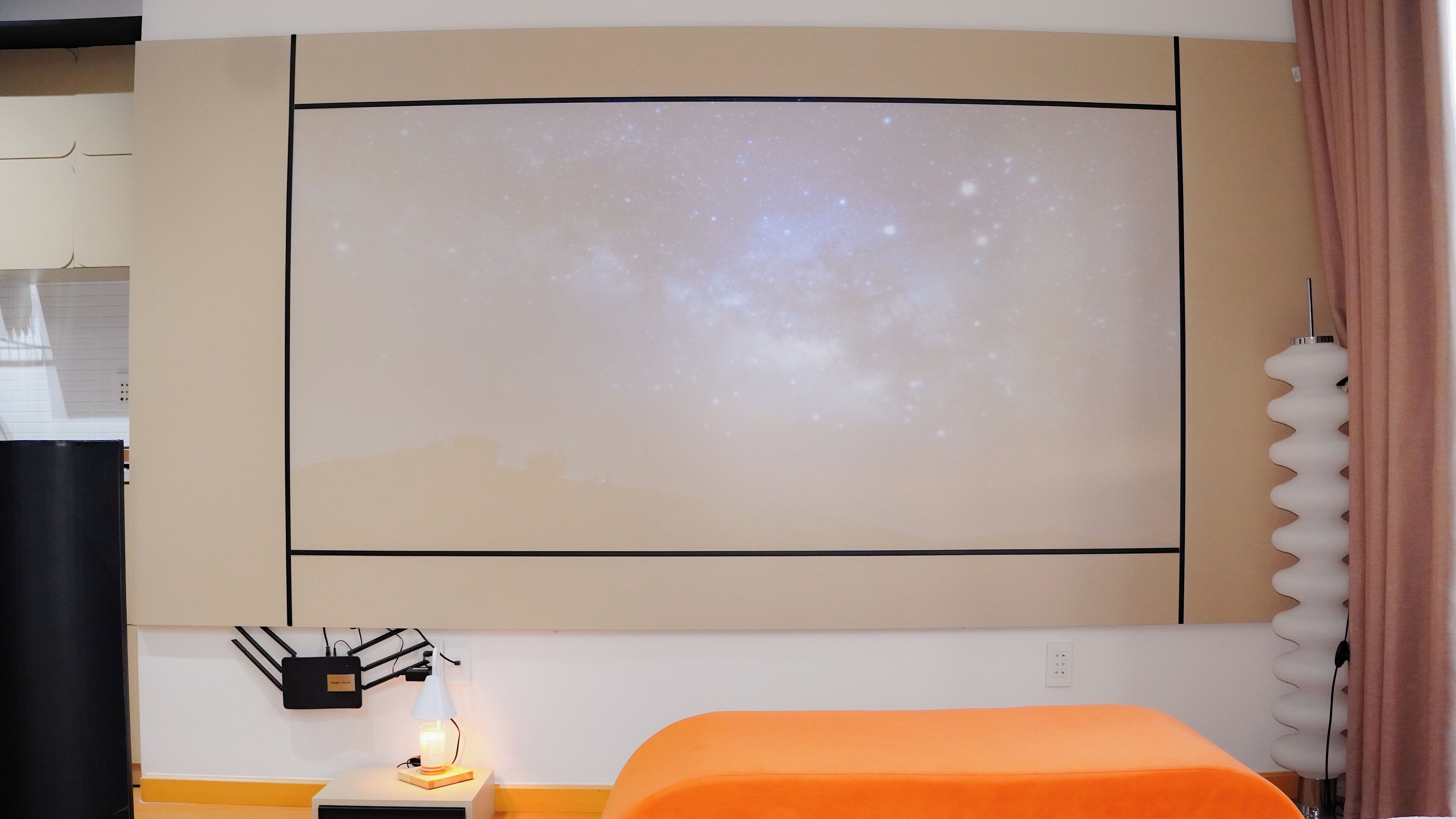 A room with a large wall-mounted projector displaying a starry sky, a bedside table with a lamp, and an orange cushioned bench.