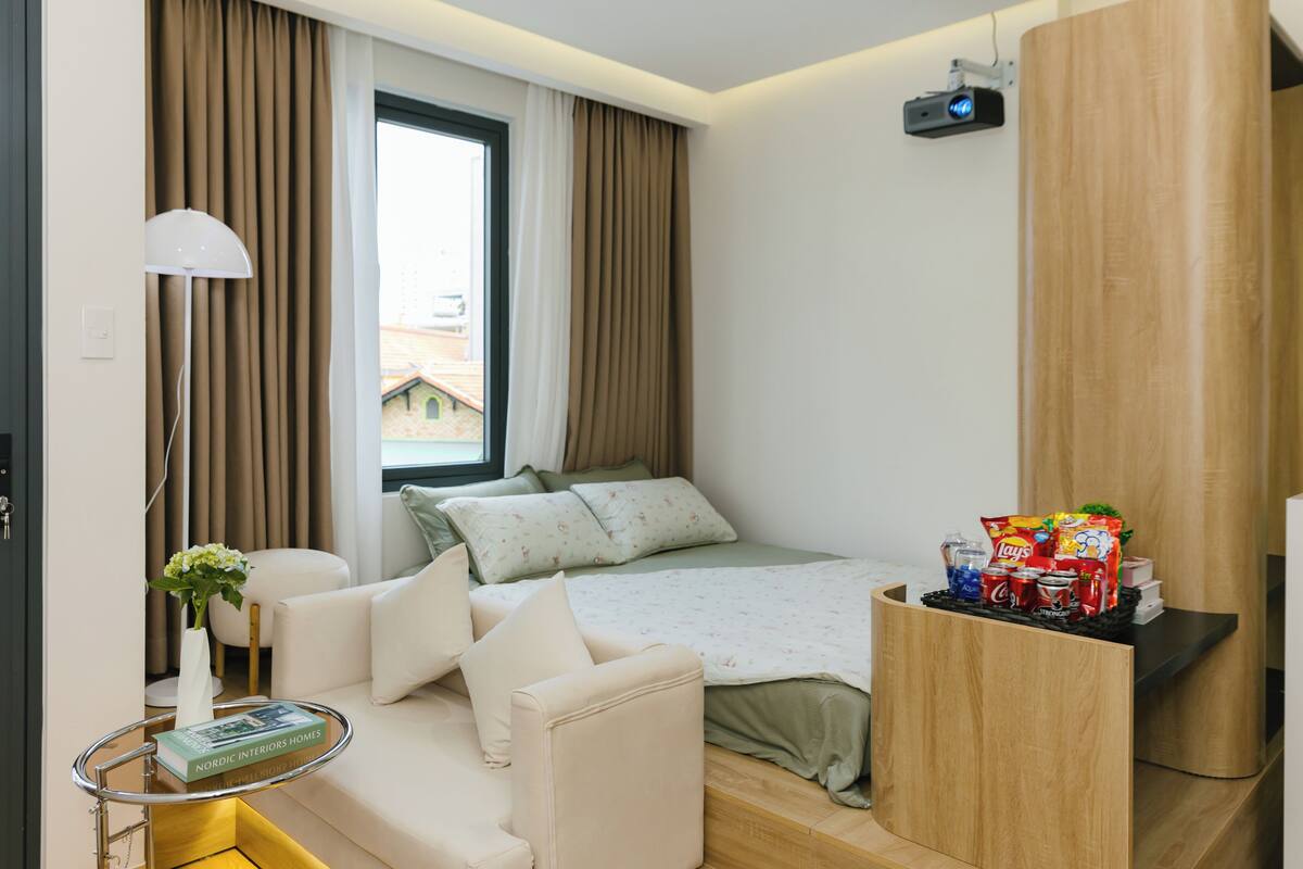 A modern hotel room with a bed, a sofa, a small table with a book, a lamp, and a window with curtains.