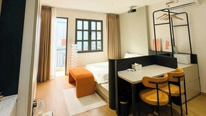 Une chambre d’hôtel moderne comprenant un lit, un bureau avec deux chaises, une petite table avec une lampe, une fenêtre avec des rideaux et une vue sur l’extérieur.