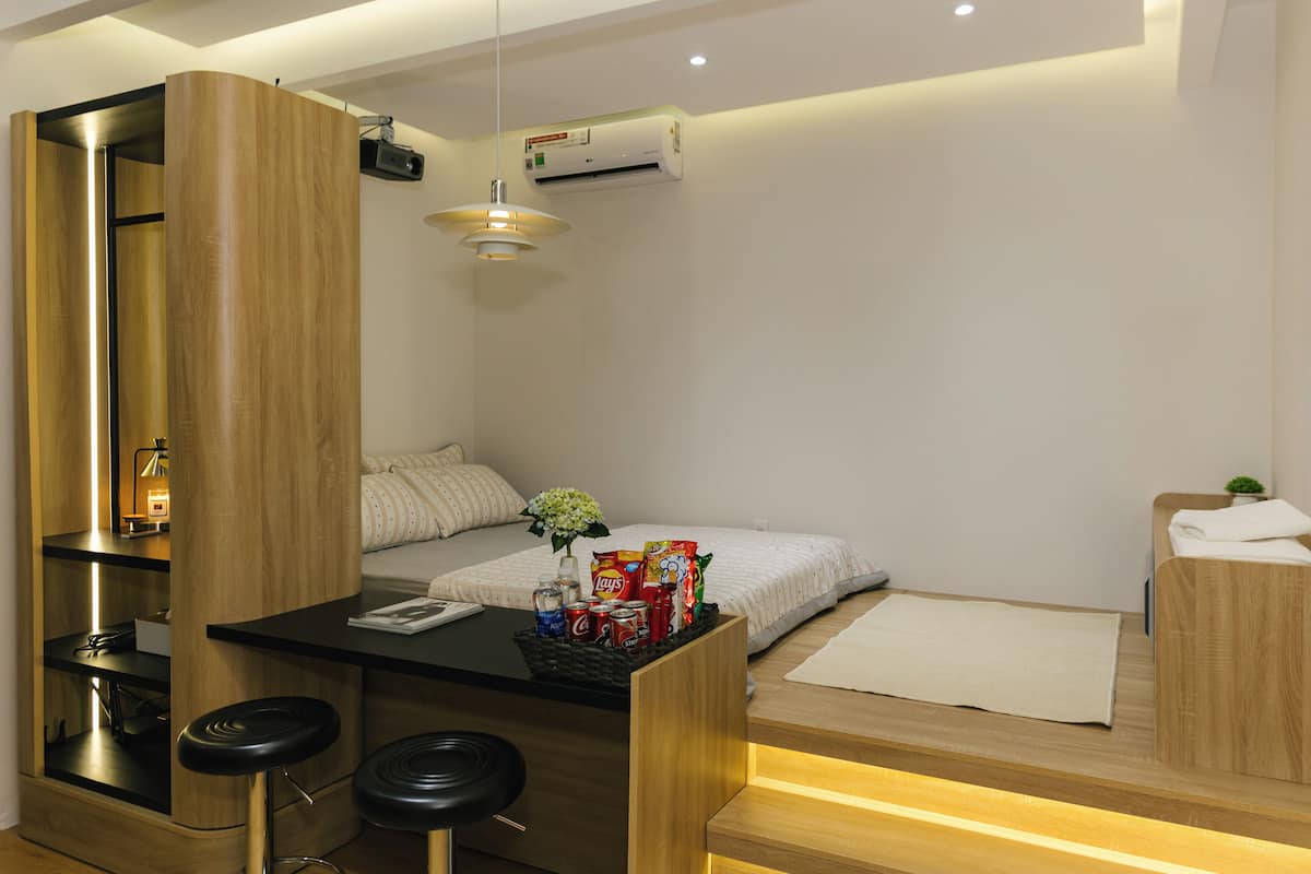 A modern hotel room with a wooden desk, bar stools, a bed with a white mattress, and a small seating area with a white rug.