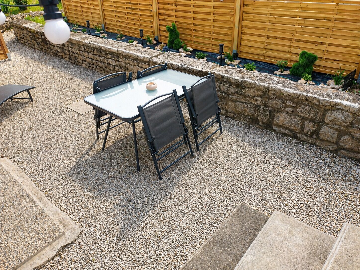 A patio with a glass table and chairs, surrounded by a stone wall and wooden fence.