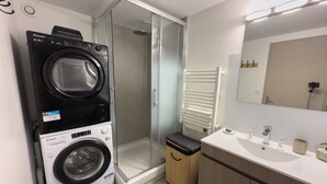 Un espace buanderie compact comprenant un lave-linge et un sèche-linge à chargement frontal, une cabine de douche en verre, un banc en bois et une salle de bain moderne avec un lavabo et un miroir.