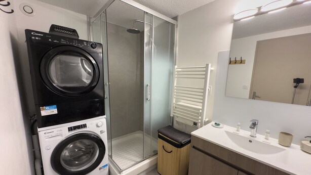 Un espace buanderie compact comprenant un lave-linge et un sèche-linge à chargement frontal, une cabine de douche en verre, un banc en bois et une salle de bain moderne avec un lavabo et un miroir.