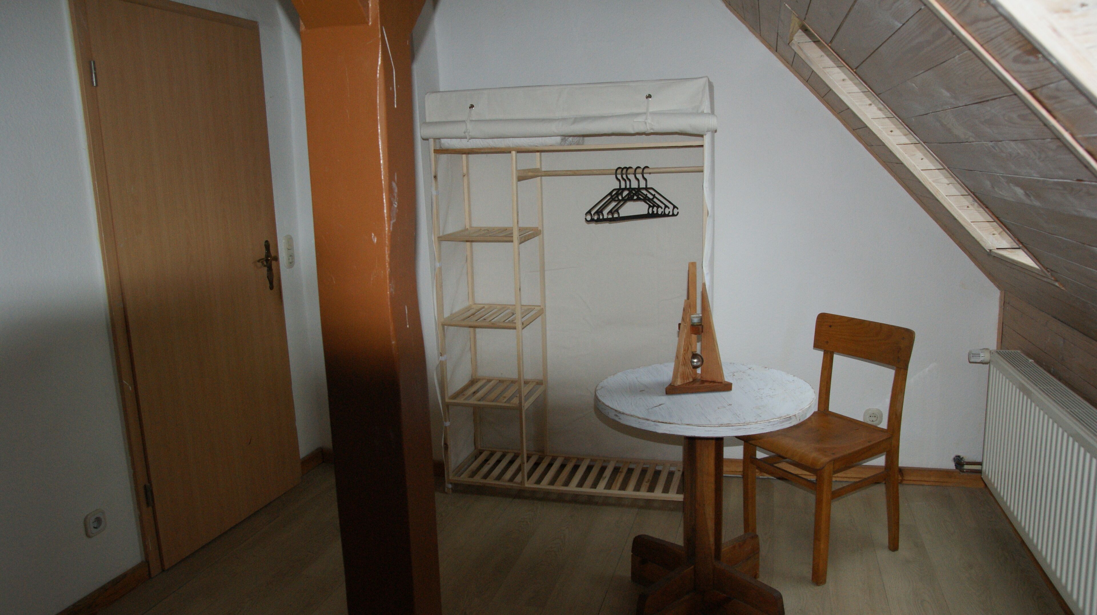 A small, cozy room with a sloped ceiling, a wooden chair, a small round table, and a white radiator.