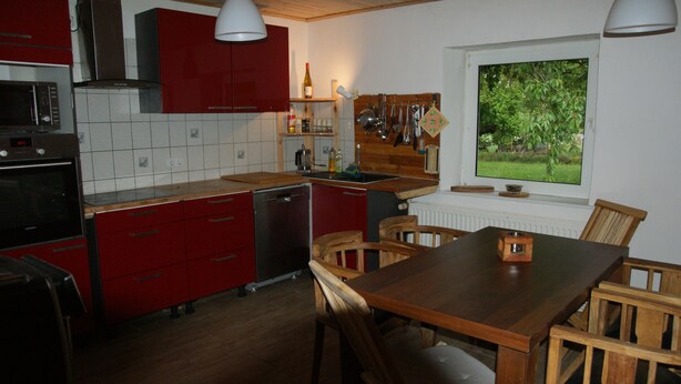 A kitchen with red cabinets, a wooden table, and a window with a view of greenery. - Farmhouse with large garden in the immediate vicinity of Lake Tollensesee (Blumenholz)