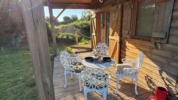 A wooden deck with white metal chairs and a table, surrounded by a wooden cabin structure. - A cottage in an orchard (Conteville)