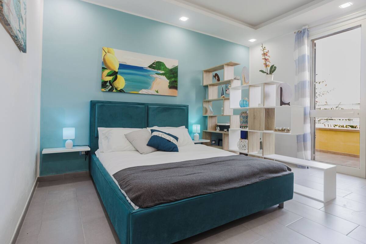 A modern bedroom with a teal bed, white nightstands, a large painting, and a wooden bookshelf.
