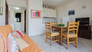 A living room with a sofa, wooden dining table, and chairs. There is a kitchen area with cabinets and a microwave. A television is on a stand in the dining area. - Lo Stagnone Apartments (Marsala)