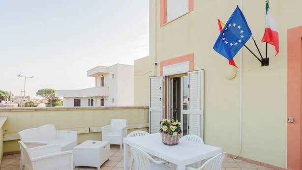 A terrace with white wicker furniture, a table with a basket of flowers, and flags on the wall. - Lo Stagnone Apartments (Marsala)