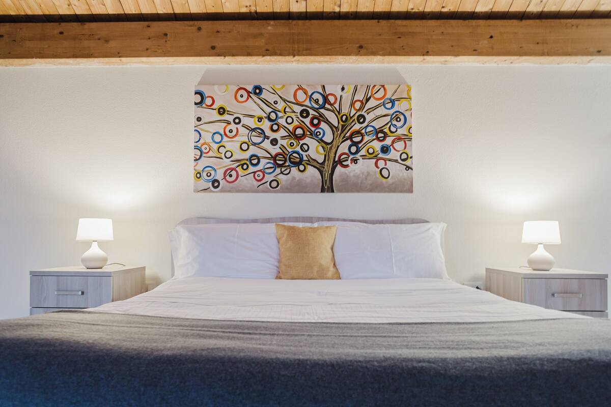 A bedroom with a bed, two bedside tables with lamps, a wall-mounted painting of a tree, and a wooden ceiling.