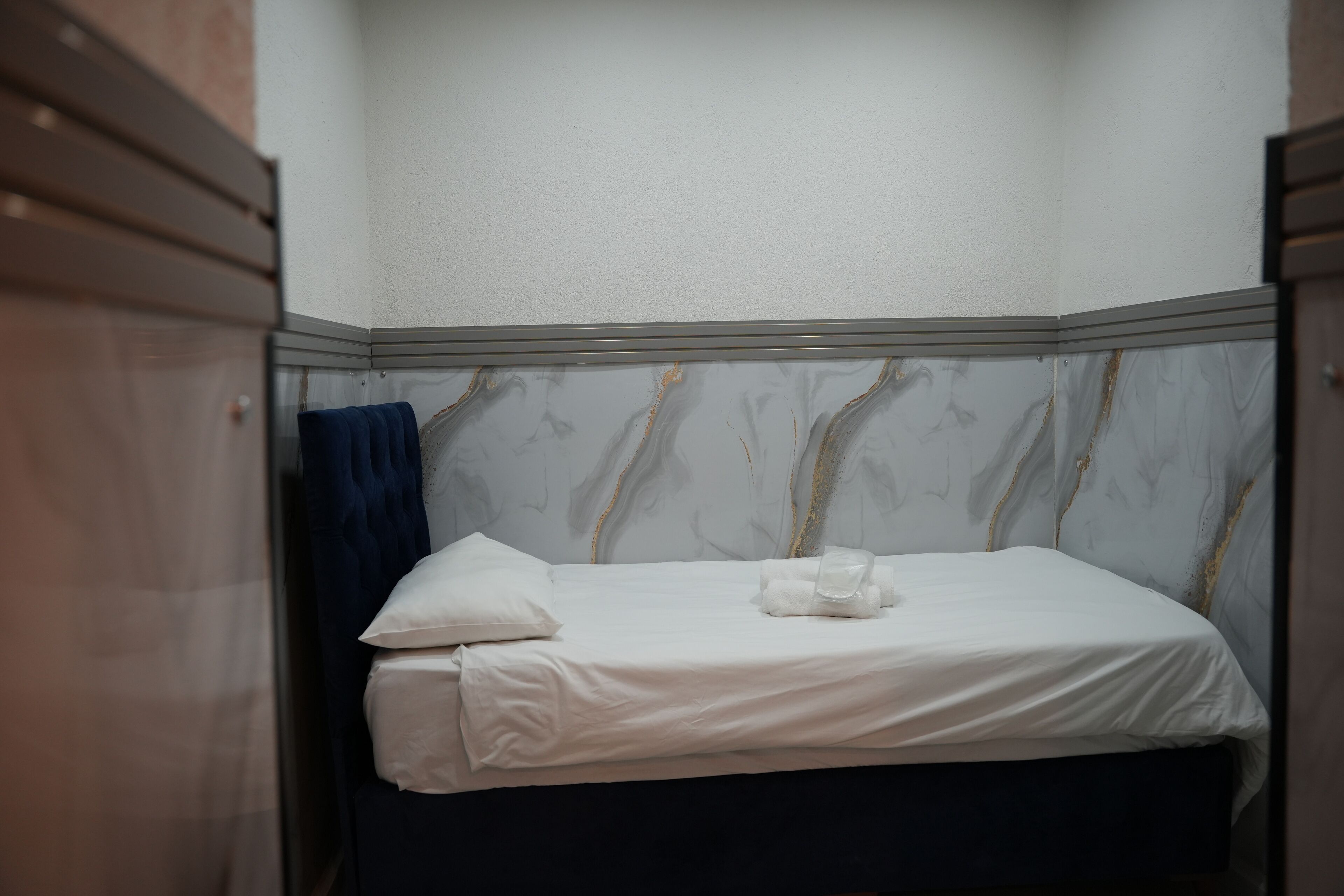 Una cama pequeña y bien hecha con sábanas blancas y una toalla azul, frente a una pared con un diseño que simula mármol.