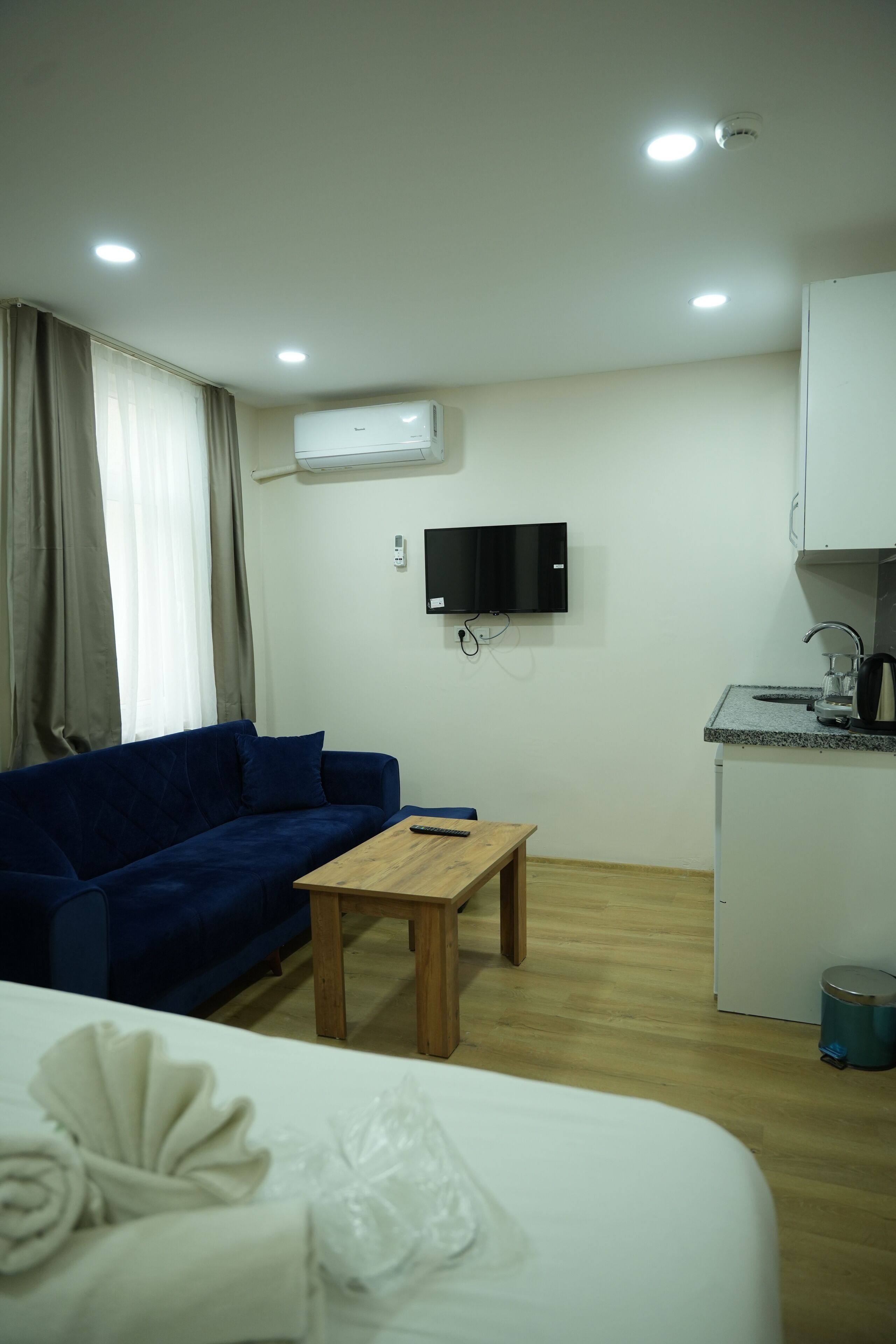 Habitación de hotel con cama, sofá azul, mesa de madera, televisor montado en la pared, pequeña cocina con fregadero y unidad de aire acondicionado.