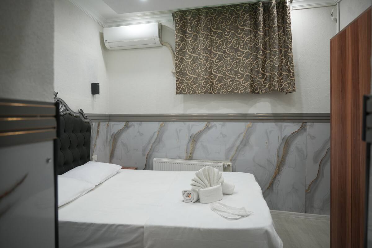A hotel room with a bed, a radiator, a wall-mounted air conditioner, and a patterned curtain.