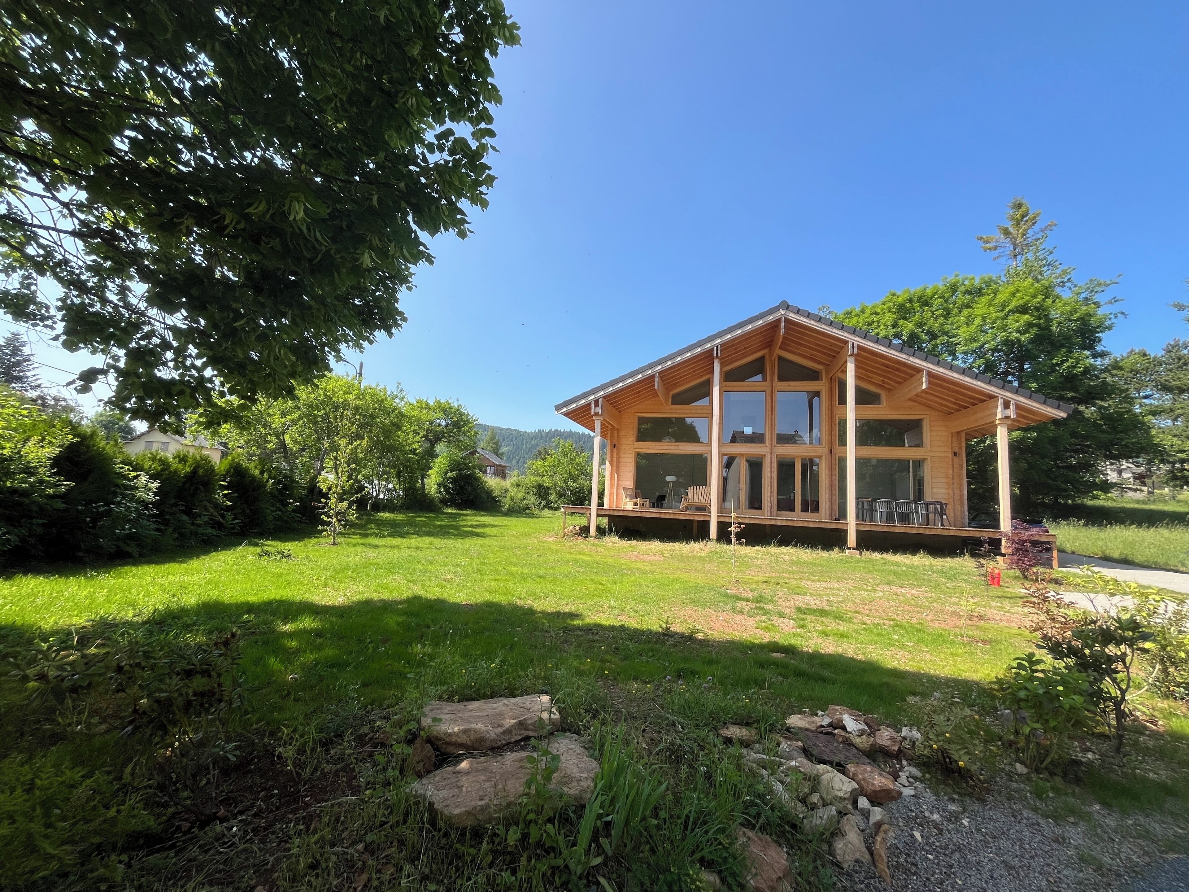 A wooden cabin with large windows, a porch with chairs, and a grassy yard.