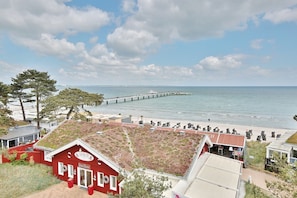 A coastal view with a pier extending into the water, a beach with sun loungers, and a red building with a green roof.