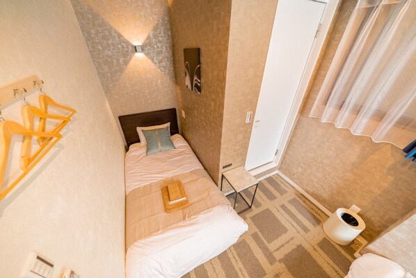 A small, compact room with a bed, a small table, a wall-mounted light, and a door.