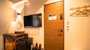 Une chambre d’hôtel compacte dotée d’une porte en bois, d’un téléviseur à écran plat, d’un bureau avec une chaise, d’un petit réfrigérateur et d’une machine à café.