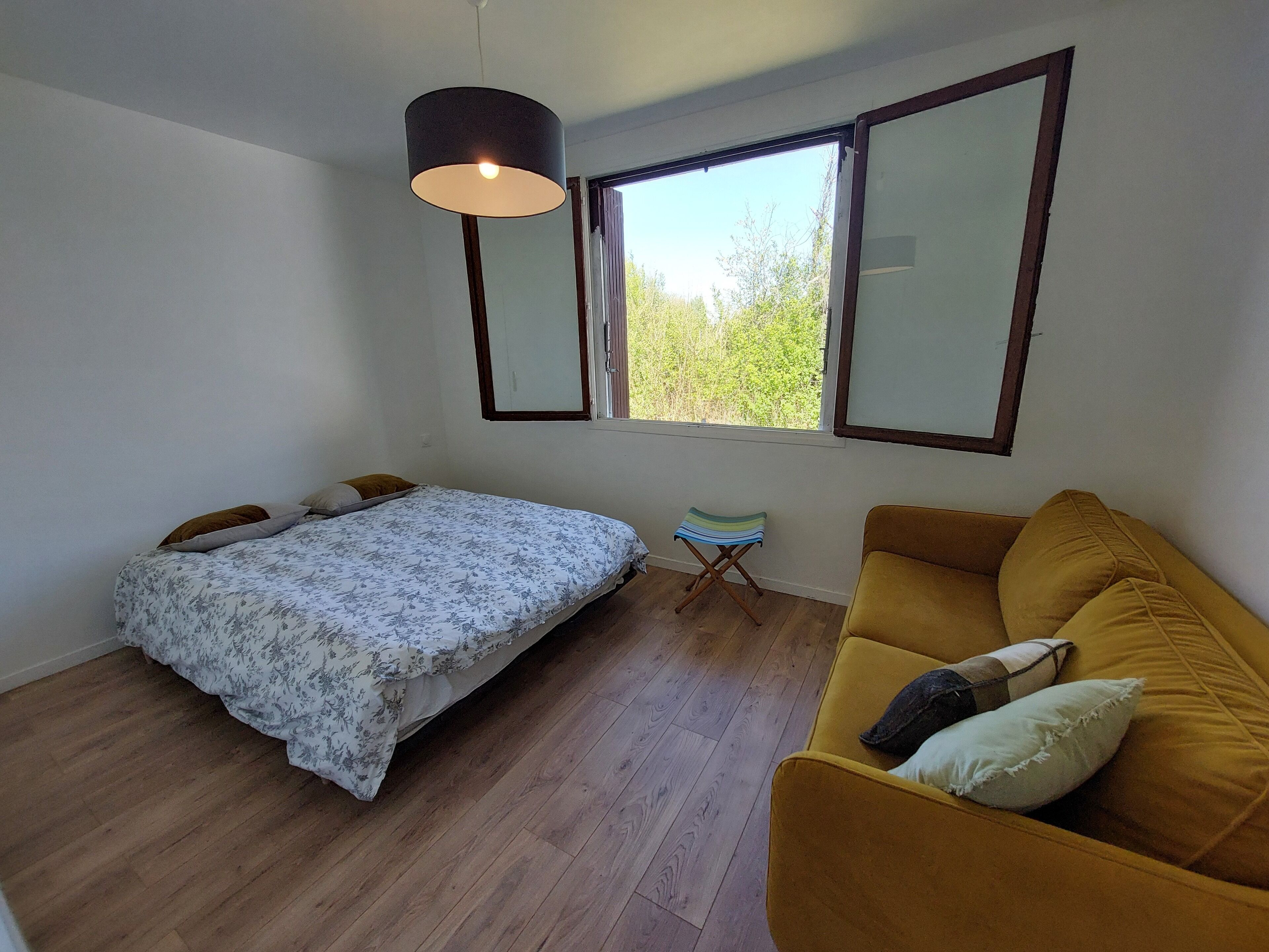 Ein Schlafzimmer mit einem Bett, einem Sofa, einem kleinen Hocker und einem Fenster mit Blick auf Grünflächen.