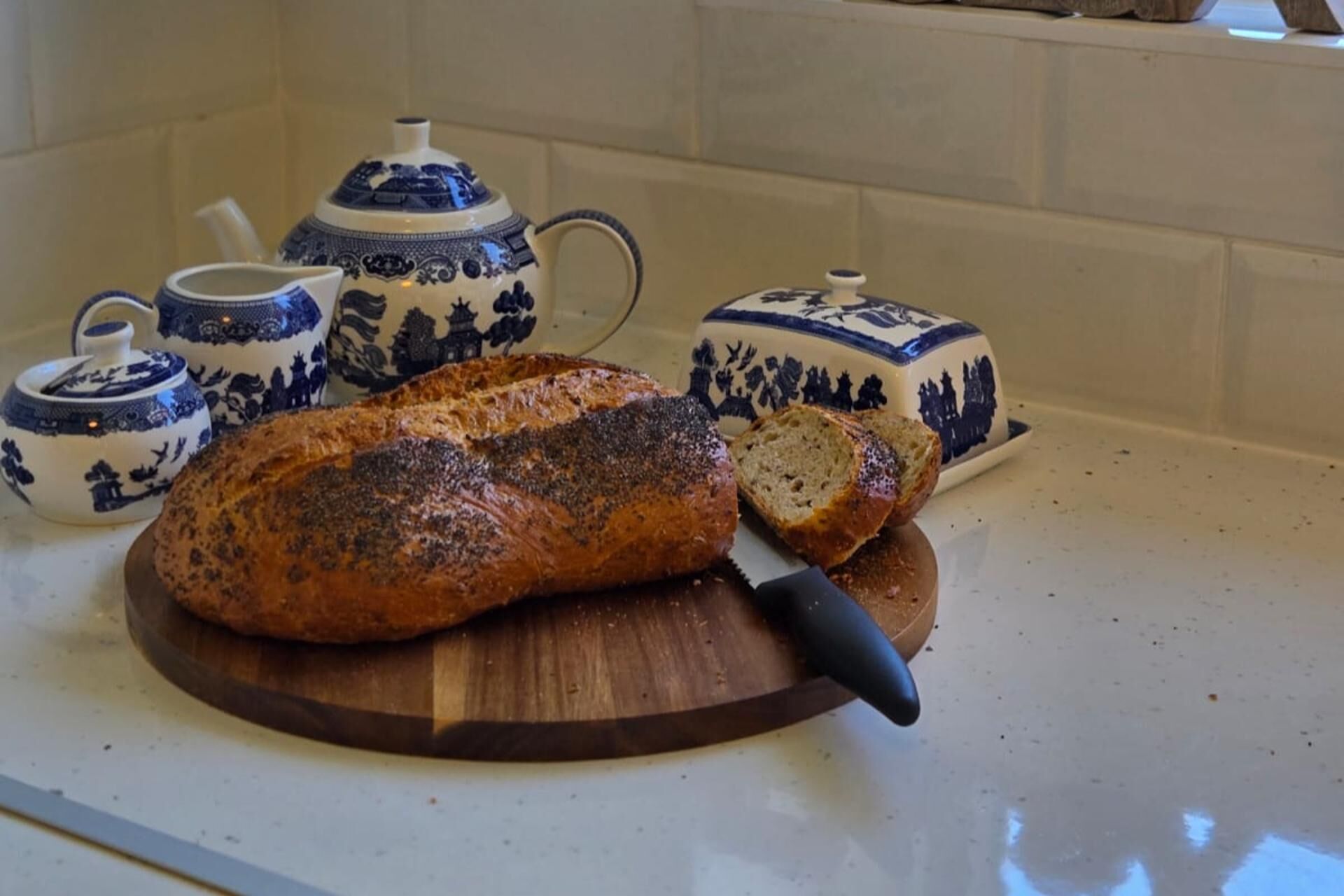 A loaf of bread on a wooden cutting board with a knife, accompanied by a teapot and two cups on a kitchen counter.