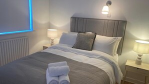 Un lit bien fait, avec une couette grise et blanche, deux lampes de chevet, une lampe fixée au mur et une fenêtre laissant filtrer une lumière bleue.