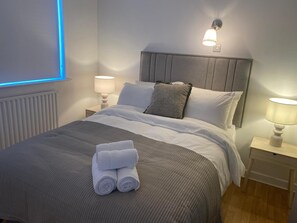 A neatly made bed with a grey headboard, white linens, and a rolled towel on the bed. There are two bedside lamps on wooden nightstands, and a wall-mounted light fixture above the bed.