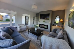A modern living room with a sofa, armchair, and a coffee table. There is a fireplace, a TV mounted on the wall, and a large mirror. - Foxes Rest - 4 Bedroom House (Tipperary)