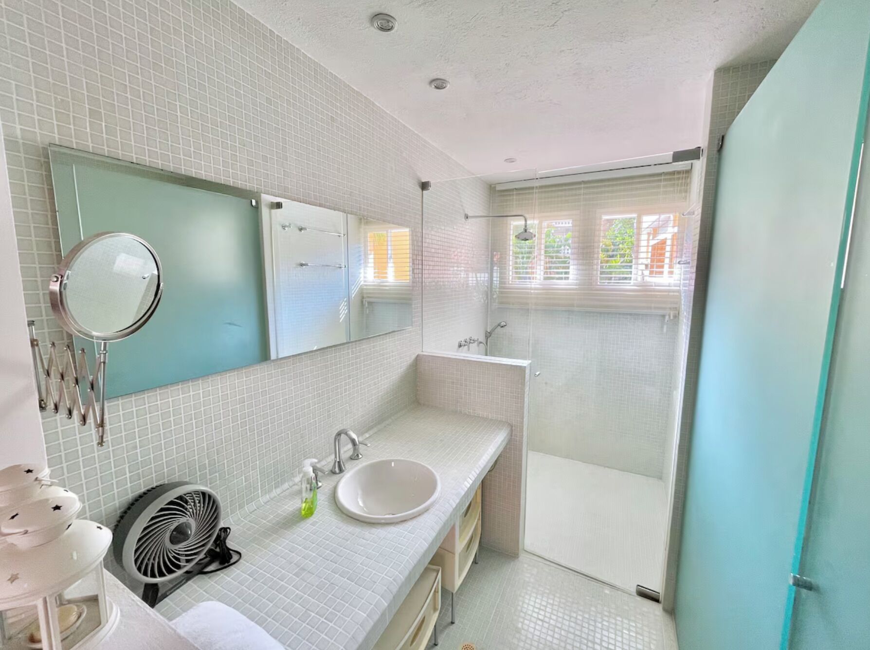 A bathroom with a white tiled wall, a round mirror, a sink, a fan, and a shower area.