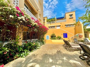 A courtyard with a tiled floor, a row of lounge chairs, and a building with balconies covered in flowering plants. - by Calamari Homes - Edificio Centenario (Cartagena)