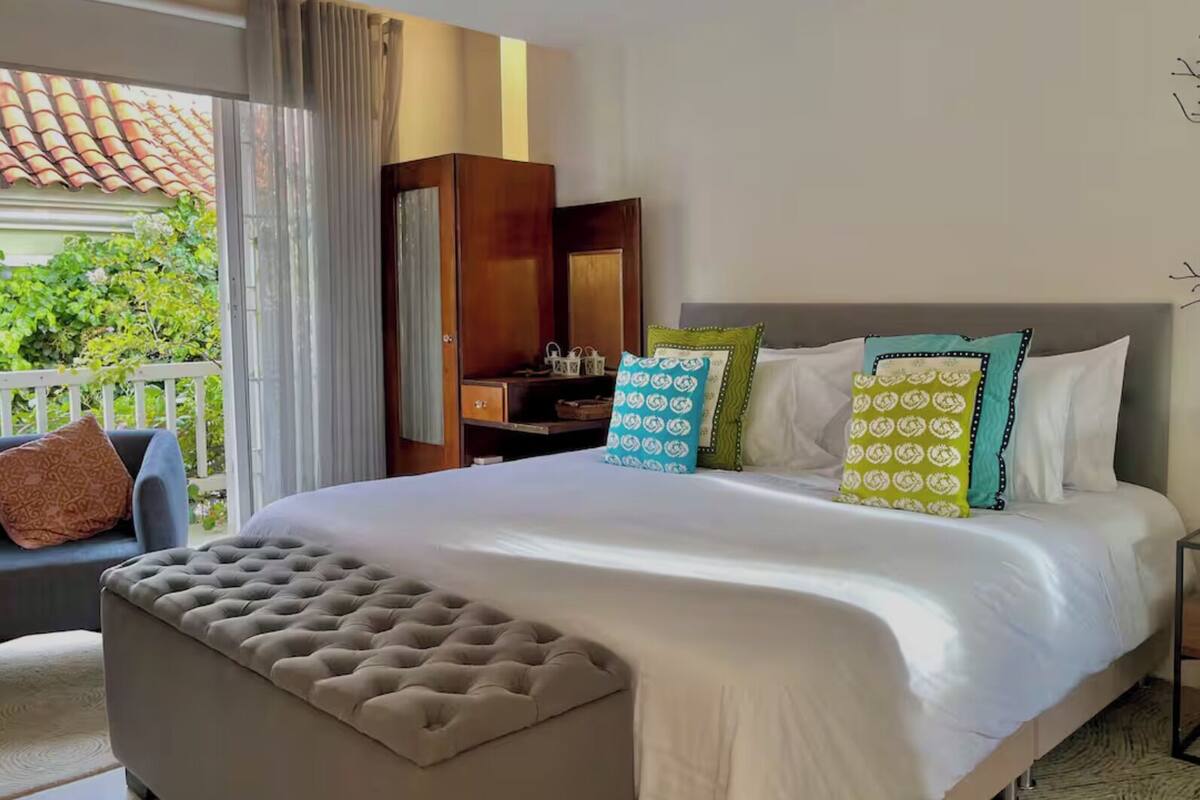 A hotel room with a large bed, a seating area with a sofa and armchair, a wooden wardrobe, and a balcony with a view of greenery.