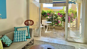 A living room with a sofa, patterned cushions, a small table, and a chair. There is a sliding glass door leading to an outdoor area with a patio and potted plants. - by Calamari Homes - Edificio Centenario (Cartagena)